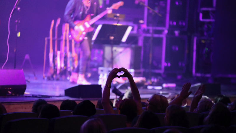 yleisön jäsen näyttää käsillään sydäntä lavan artisteille hämärässä konserttisalissa
