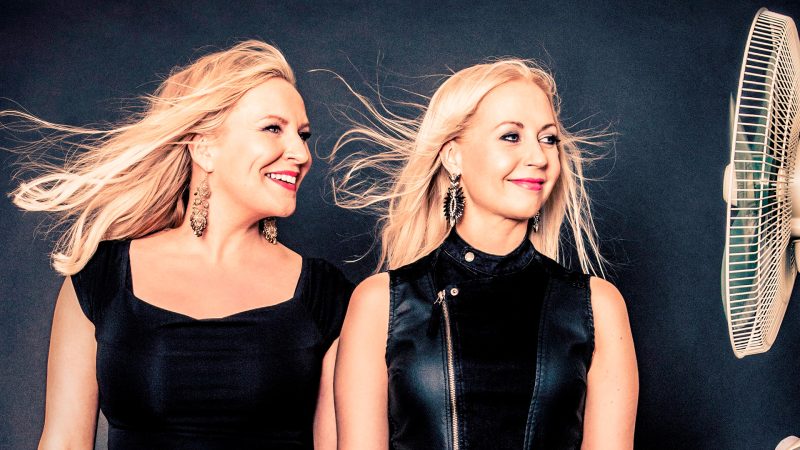 Kaksi blondia naishahmoa katsovat oikealle ja tuulikone puhaltaa heidän hiuksiinsa.