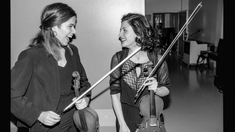 Kaksi viuslistinaista keskustelevat viulut kädessään mustavalkoisessa kuvassa.