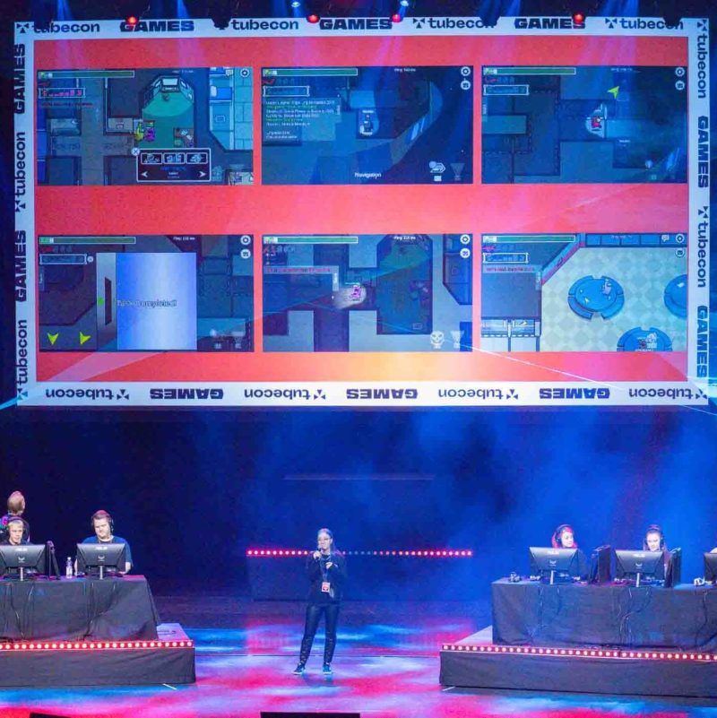 Tubecon Games pelitapahtuma Tampere-talon Isossa salissa, jossa juontaja selostaa lavalla pelaajien istuessa pelikoneiden ääressä lavan sivuilla