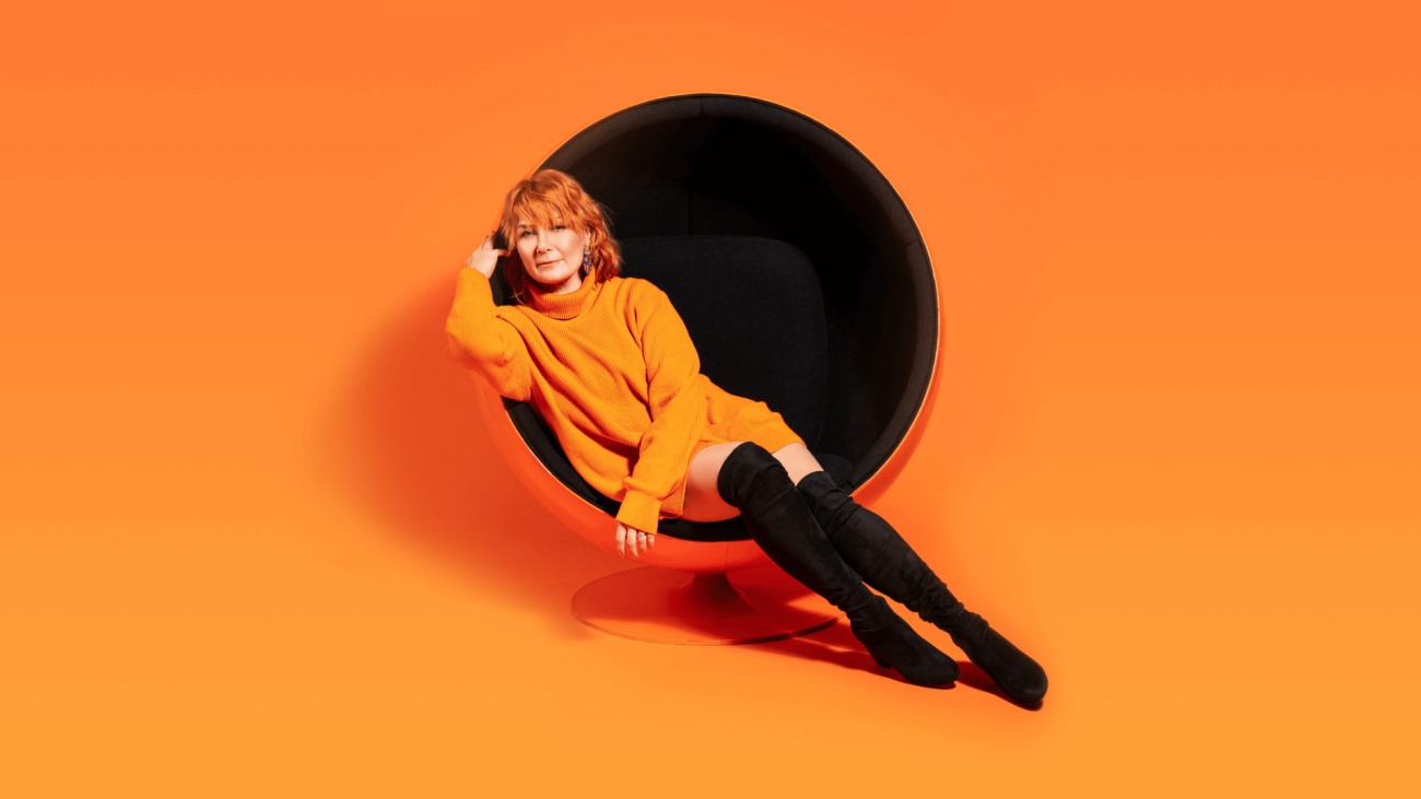 Artisti Irina oranssissa tukassa, oranssissa paidassa, oranssin taustan edessä