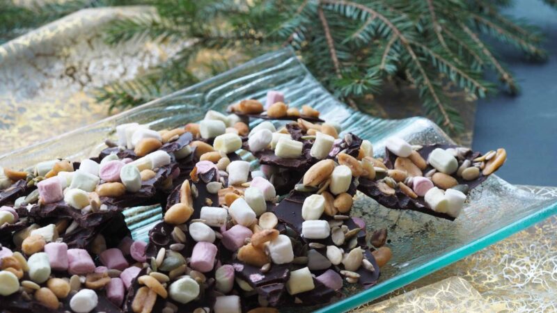 Kuvassa lasiastialla jouluinen Rocky road -jälkiruoka, jossa on suklaata, vaahtokarkkeja, pähkinää sekä siemeniä. Kuvassa dekoraationa kultainen pöytäliina sekä havunoksia.