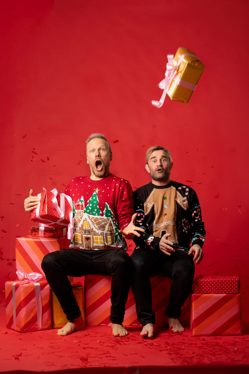 Kaksi joulupaitaista miestä istuvat punaisessa tilassa lahjapakettien päällä