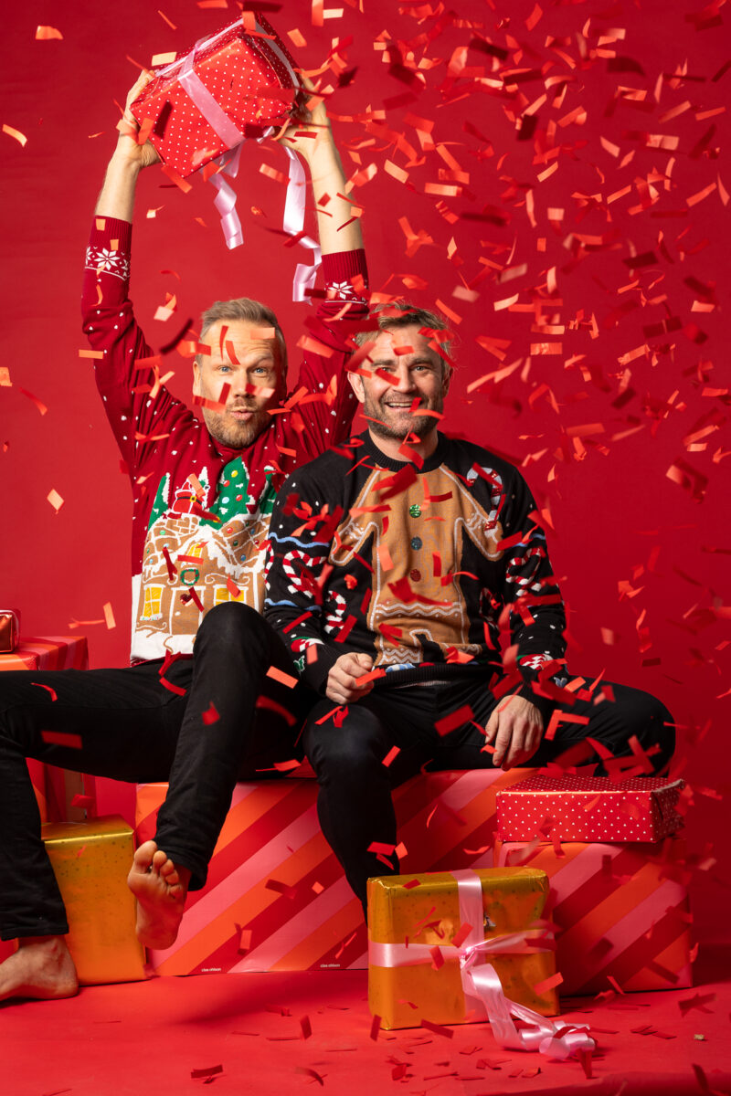 Kaksi joulupaitaista miestä istuvat punaisessa tilassa lahjapakettien päällä