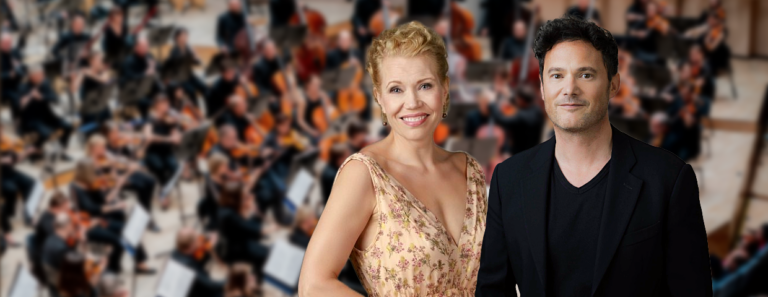 Kapellimestari ja sopraano etualalla ja orkesteri sumeana taustalla.
